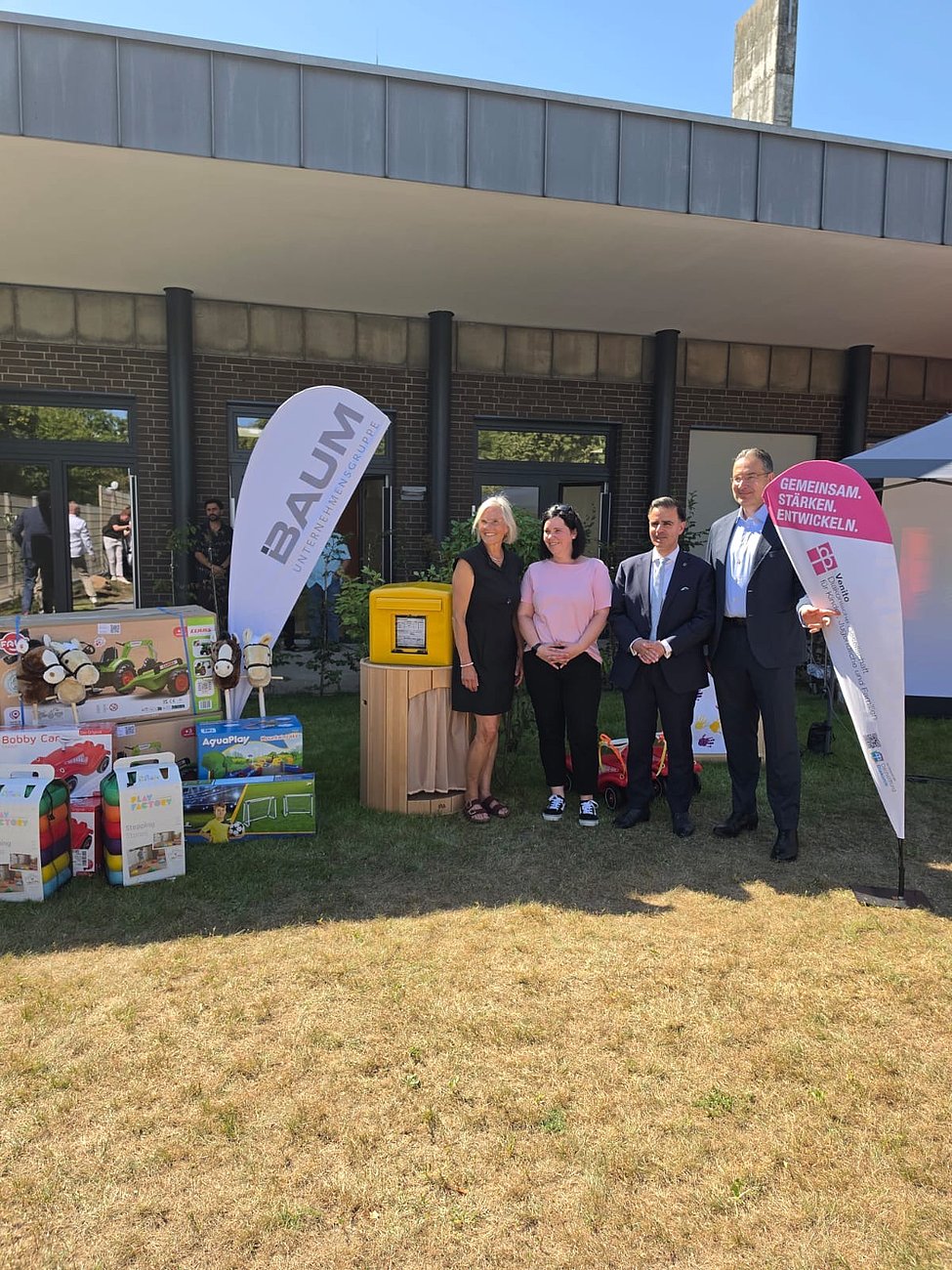 Vier Personen vor Infostand mit Werbefahnen, Produktpaketen und moderner Gebäudefassade; sonniger Tag auf trockenem Rasen.
